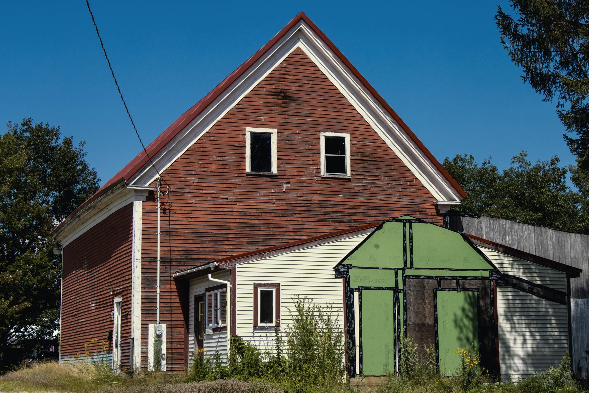A gable-end farmhouse mid-residing, new house-wrap and boards going up beside the old red siding