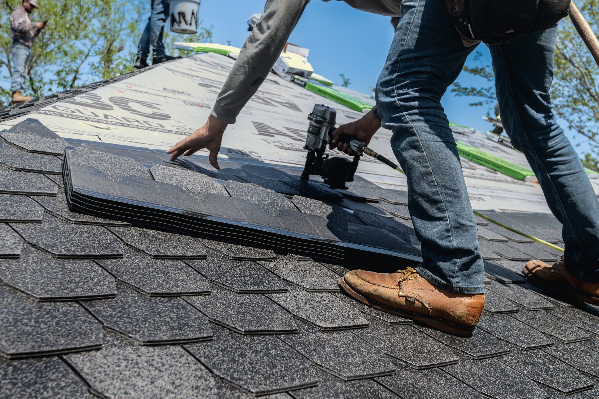 A Halden roofer nailing off a course of architectural shingle, worn work boots braced on the slope